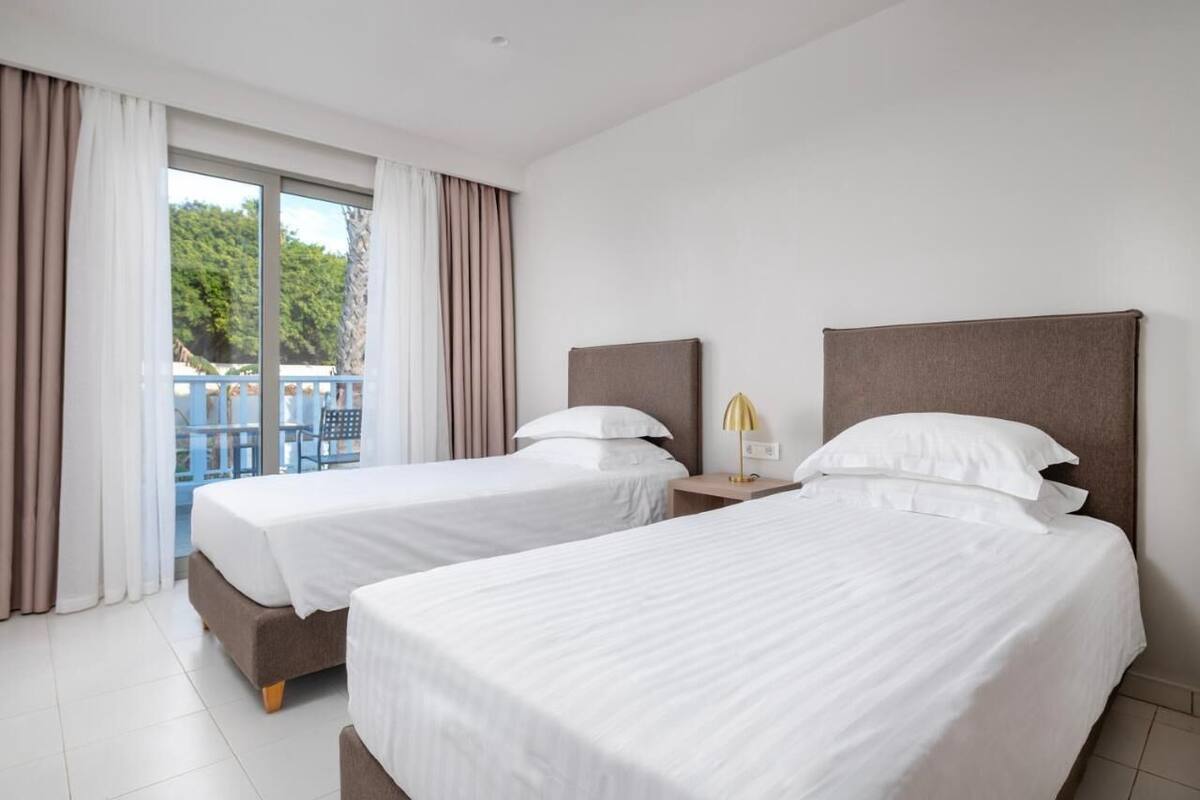 A hotel room with two beds, a balcony with curtains, and a view of greenery outside.