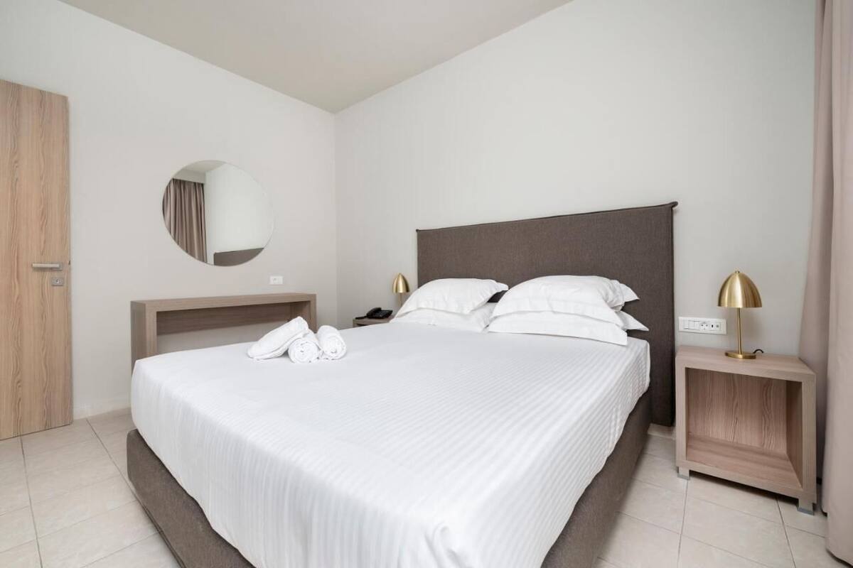 A modern hotel room with a large bed, a round mirror, a bedside table with a lamp, and a wooden wardrobe.