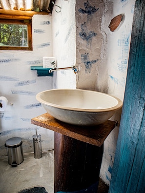 A bathroom with a large, round, beige sink mounted on a wooden pedestal. There is a small shelf above the sink holding a roll of toilet paper. The walls are partially painted with blue patterns, and there is a window with a view of trees outside.