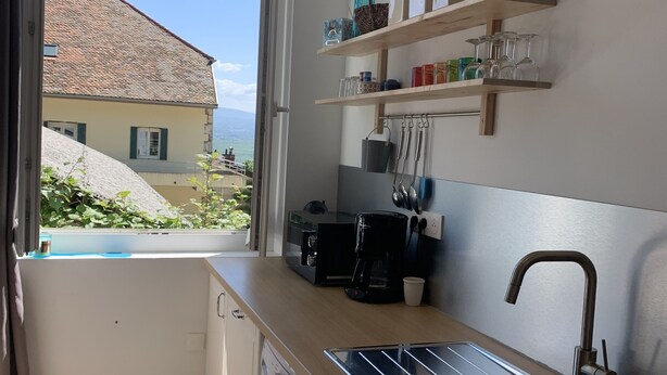 Cocina compacta con lavadora, cafetera y lavamanos. Hay estantes con frascos y vasos. Una ventana ofrece vistas a una casa y follaje.