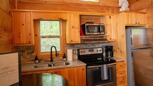 A wooden cabin kitchen with a stone fireplace, a stainless steel refrigerator, a microwave, a stove, and a sink. - Serenity Ridge Cabin E: Couple's Holiday (Mill Spring)