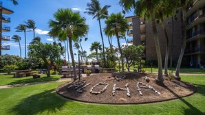 A landscaped area with a circular stone arrangement spelling 'LOHA' surrounded by palm trees and a building with balconies. - Modern and Relaxing Oceanfront Vistas (Kihei)