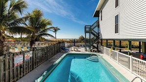 A pool area with a wooden deck, palm trees, and a white building with a balcony.