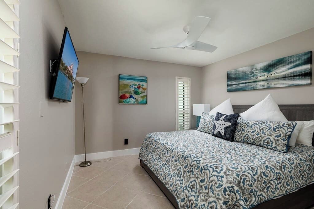 A bedroom with a bed, a ceiling fan, a TV, a floor lamp, and artwork on the walls.