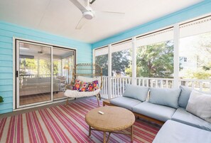A covered porch with a sofa, a wicker table, and a chair with colorful cushions. - Tropicana Cabana with Fenced Yard (Isle Of Palms)