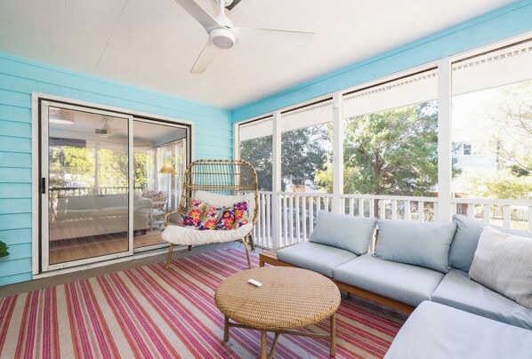 A covered porch with a sofa, a wicker table, and a chair with colorful cushions. - Tropicana Cabana with Fenced Yard (Isle Of Palms)