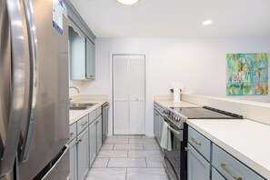 A modern kitchen with stainless steel appliances, a white countertop, and a colorful abstract painting on the wall. - Tropicana Cabana with Fenced Yard (Isle Of Palms)