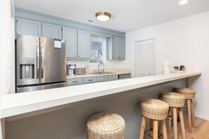 Une cuisine moderne avec des appareils en acier inoxydable, des meubles de couleur bleu clair et un plan de travail blanc. On y trouve trois tabourets en osier et une fenêtre qui laisse entrer la lumière naturelle.