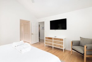 Une chambre à coucher avec un lit, une table de chevet blanche dotée de tiroirs en osier, un canapé gris, un téléviseur fixé au mur et une porte donnant sur une salle de bain.