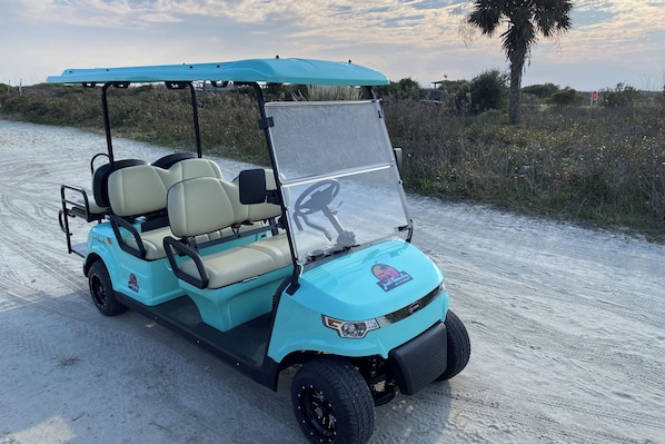 A golf cart parked on a sandy path with a blue canopy, beige seats, and a steering wheel.