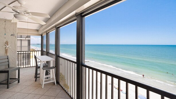 A balcony with a table and chairs, a ceiling fan, and a view of the beach and ocean.