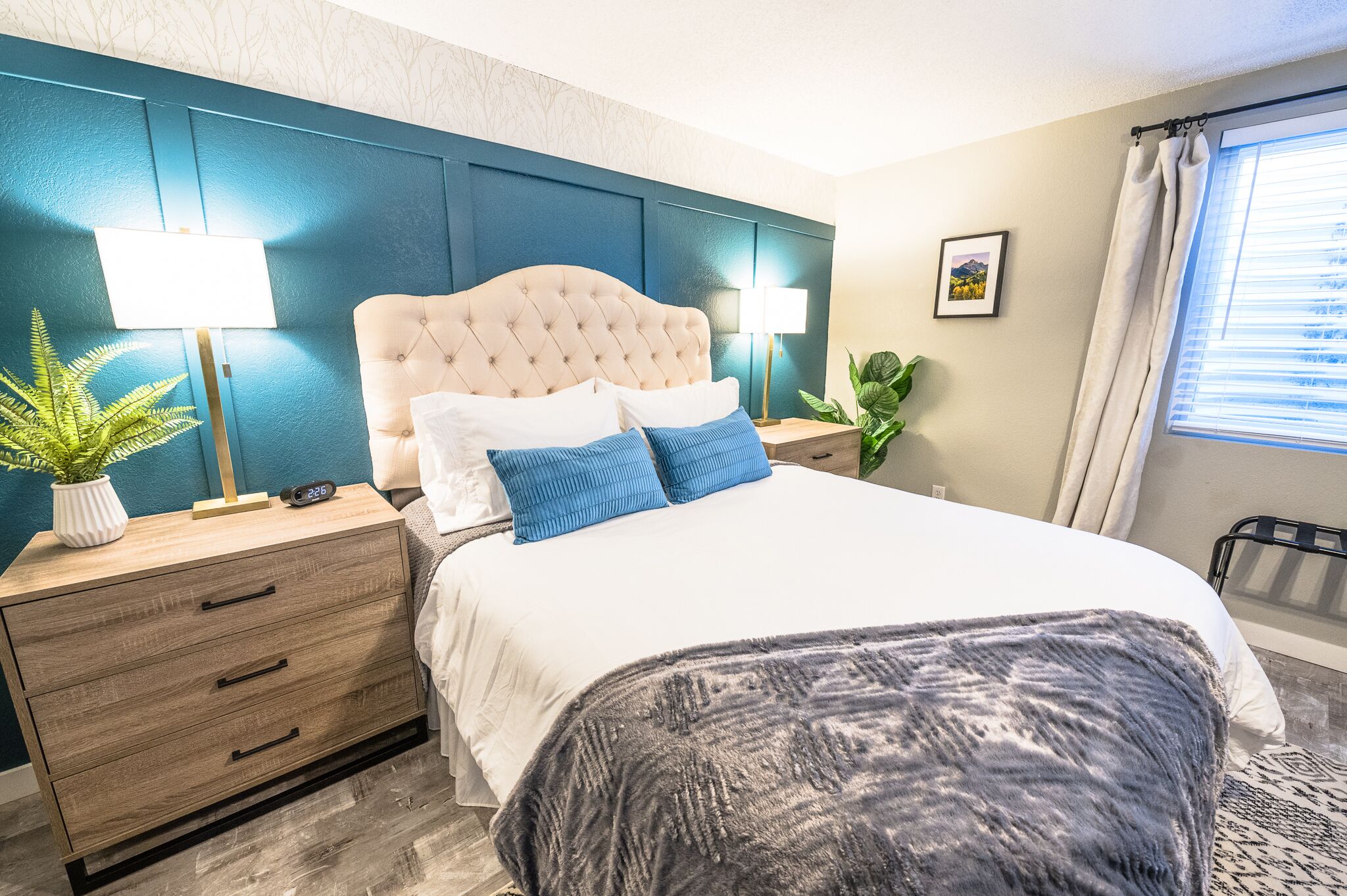 A bedroom with a tufted headboard, a white bedspread, blue and white pillows, a grey throw blanket, a bedside table with a lamp, a clock, and a potted plant.