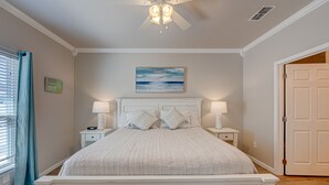 Une chambre avec un grand lit, deux tables de chevet avec des lampes, un ventilateur de plafond, un tableau au mur et une porte sur le côté droit.