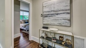 A hallway with a modern console table, a large abstract painting, and a glimpse of a bedroom with a bed and window. - Cozy Riverside Villa - Walk to Bars & Restaurants! (Jacksonville)