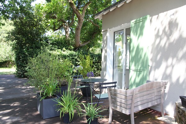 A patio with a table, chairs, and a bench. There are potted plants and a tree in the background.