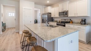 A modern kitchen with white cabinets, a granite countertop, stainless steel appliances, and wooden bar stools. - Bring The Whole Family And Friends, Too! White Beaches Are Waiting! (Perdido Key)