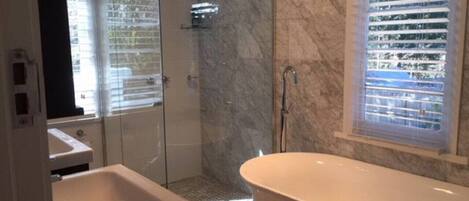 Un baño con ducha envuelta en vidrio, una bañera independiente y una ventana con persianas.