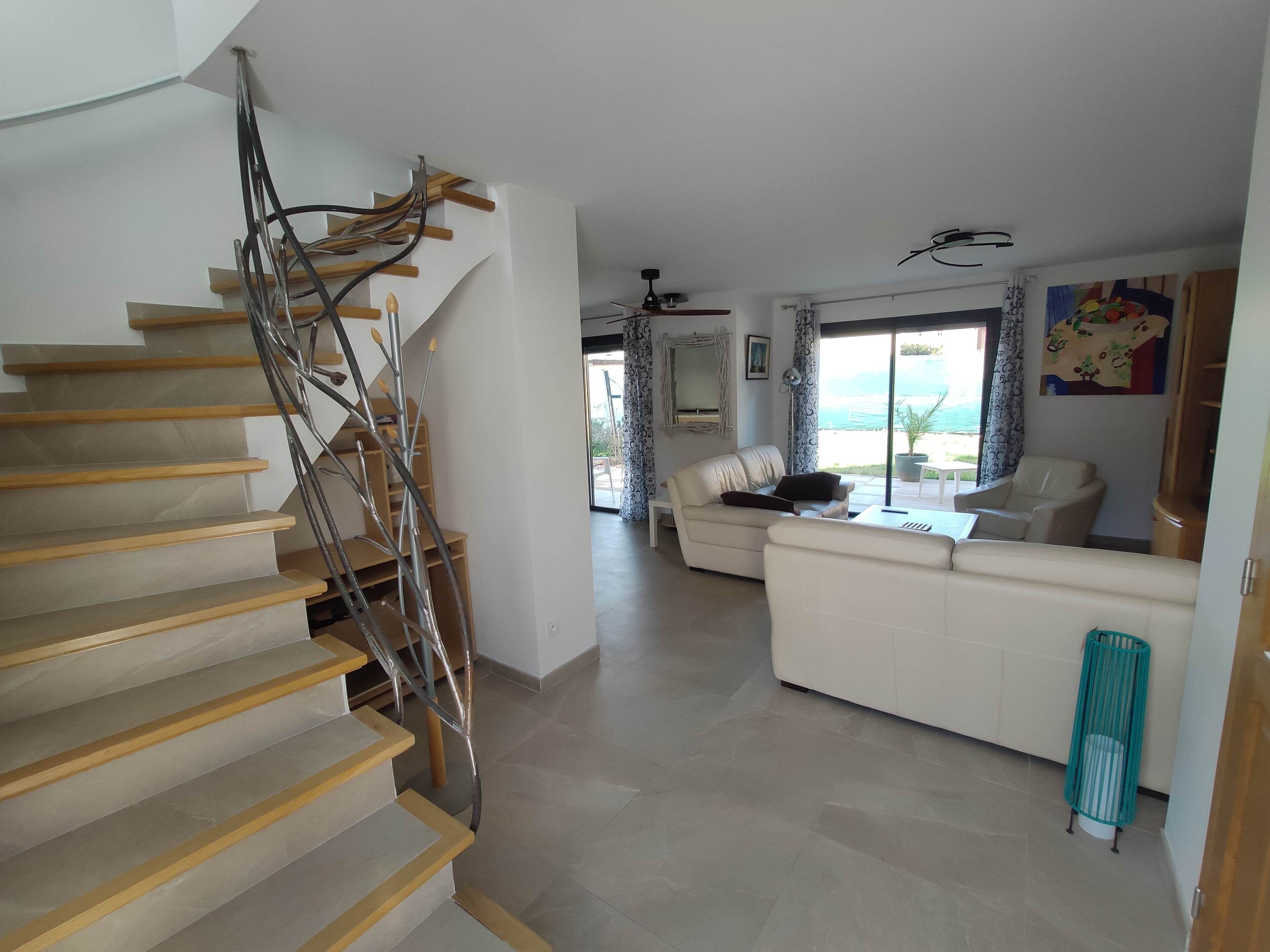 Un escalier avec des marches en bois et des rampes en métal, menant à un espace de vie comprenant un canapé blanc, une table basse et une vue sur l’extérieur.