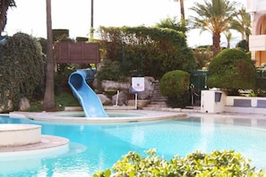 Une piscine avec un toboggan bleu, entourée d’un aménagement paysager soigné et de palmiers.