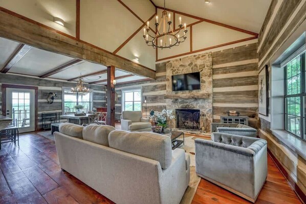 A spacious living room with a stone fireplace, large windows, and a chandelier.