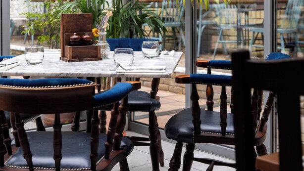 An indoor dining area with wooden chairs and tables, large windows, and potted plants. - The Pilot (London)