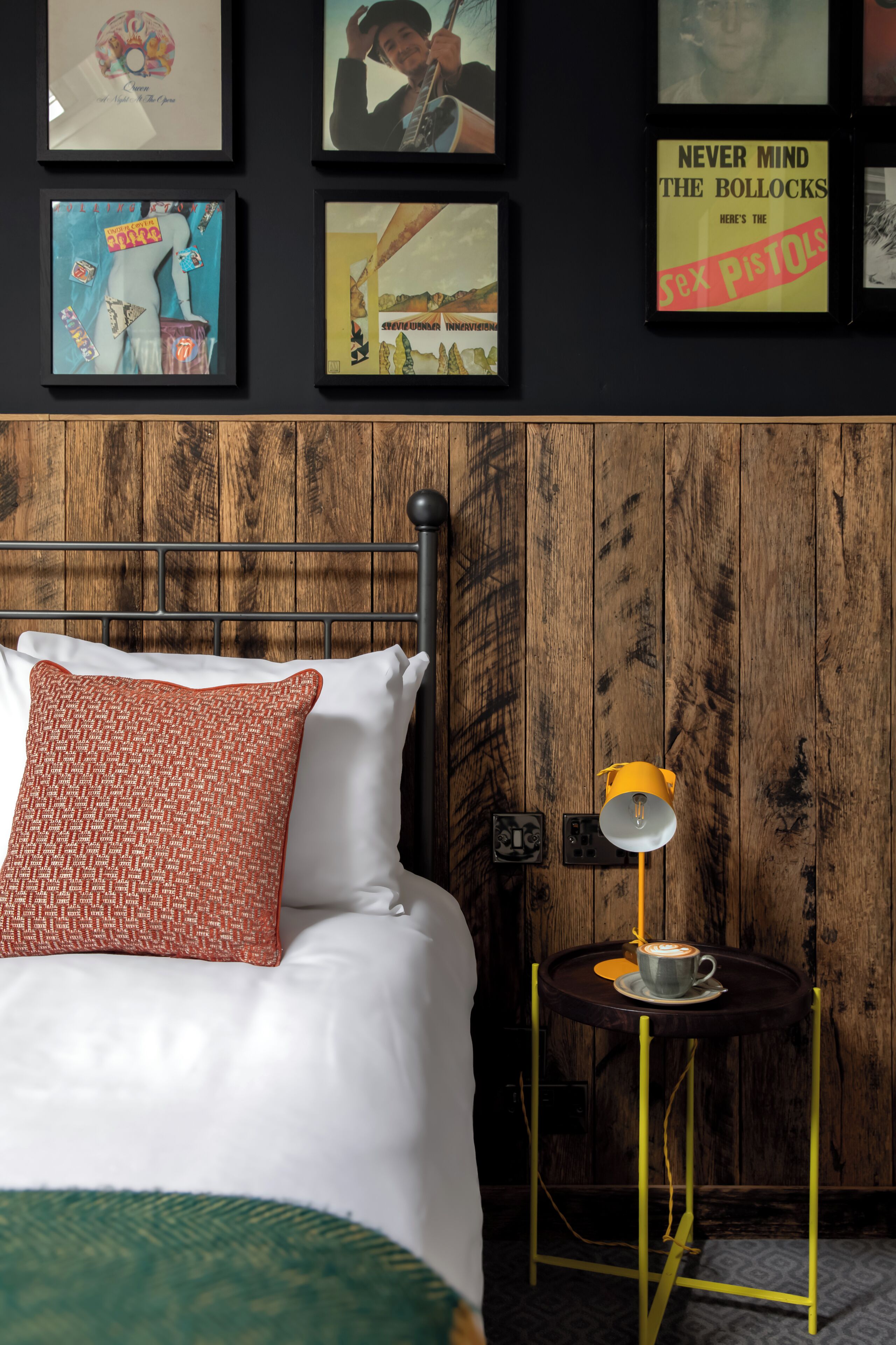 A bedroom with a wooden headboard, a bed with white bedding and a patterned pillow, a small table with a cup and saucer, and a wall with framed pictures.
