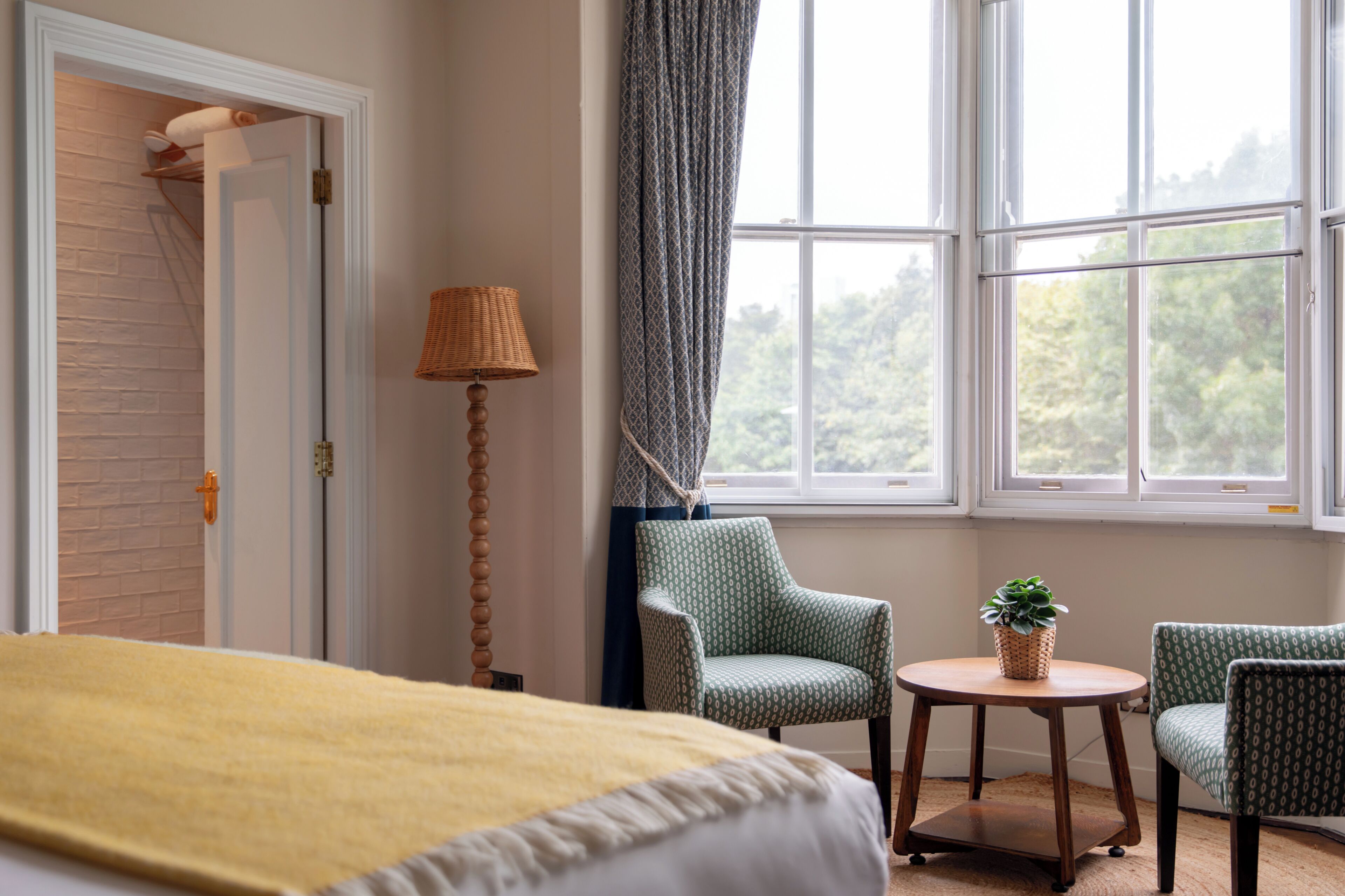 A hotel room with a bed, two chairs, a small table with a plant, a lamp, and a window with a view of greenery.