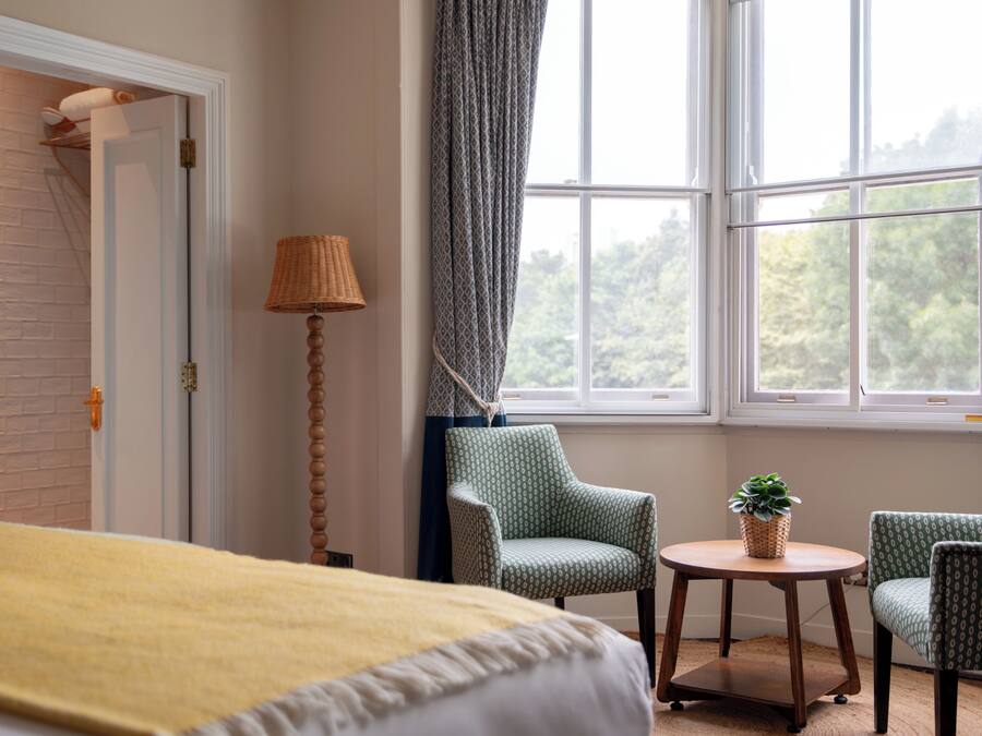 A hotel room with a bed, two chairs, a small table with a plant, a lamp, and a window with a view of greenery.