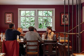 A dining area with tables and chairs, a window with a view of greenery, and framed pictures on the wall.