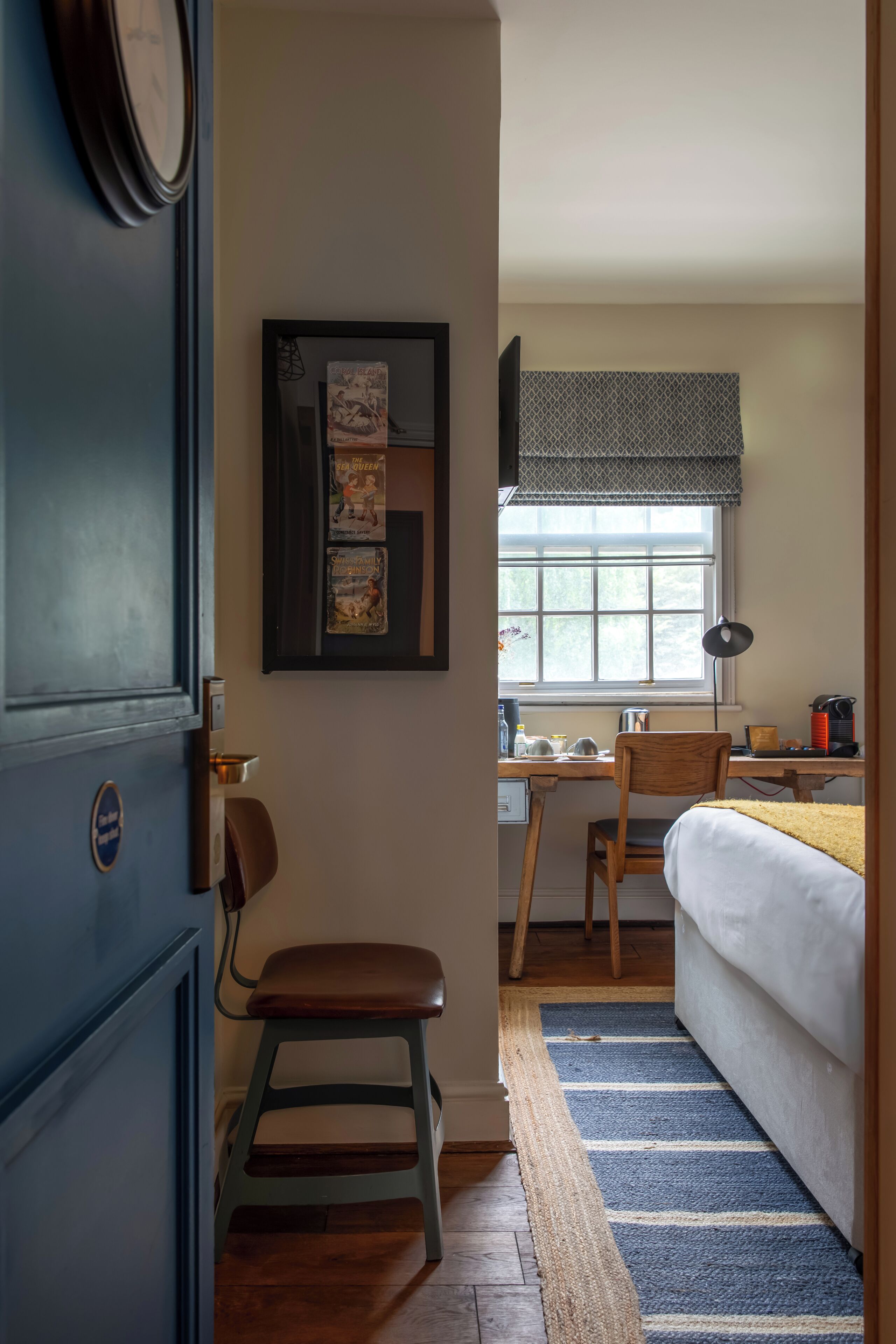 Habitación de hotel pequeña con cama, escritorio, silla, taburete, reloj y ventana con vistas.