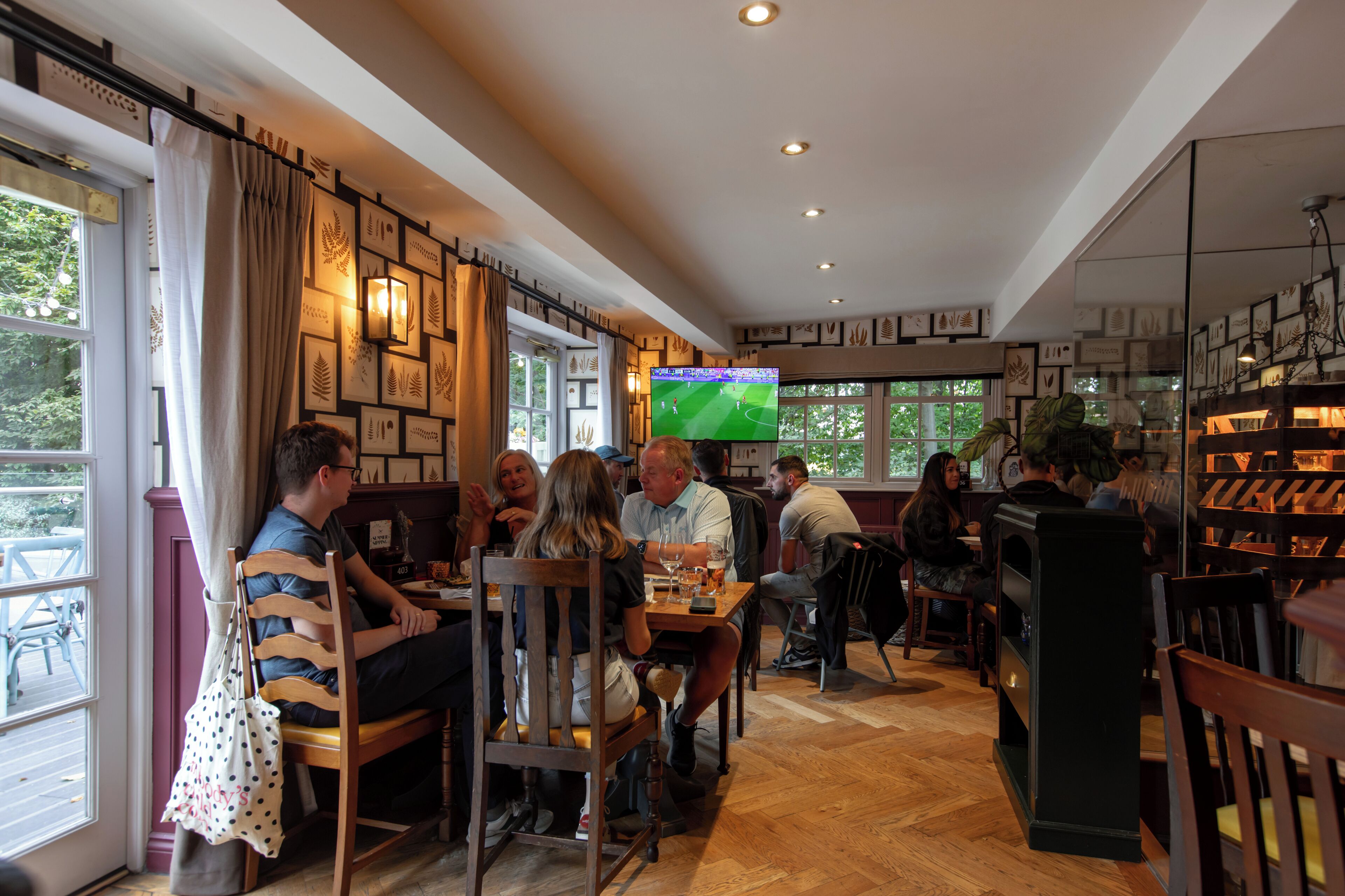 A restaurant with wooden furniture, a TV screen displaying a sports game, and framed botanical prints on the walls.