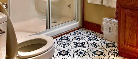 Un baño con ducha de vidrio, inodoro, lavabo con gabinete de madera, pisos de baldosas estampadas y decoración en la pared.