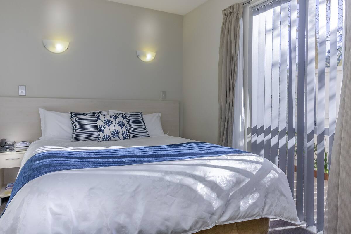 Een netjes opgemaakt bed met een wit-blauwe streepdeken, een wit kussen met een blauw-wit patroon, en een effen wit kussen. Er staat een nachtkastje met een lamp en telefoon op. De kamer heeft een raam met dunne gordijnen en uitzicht op bomen.