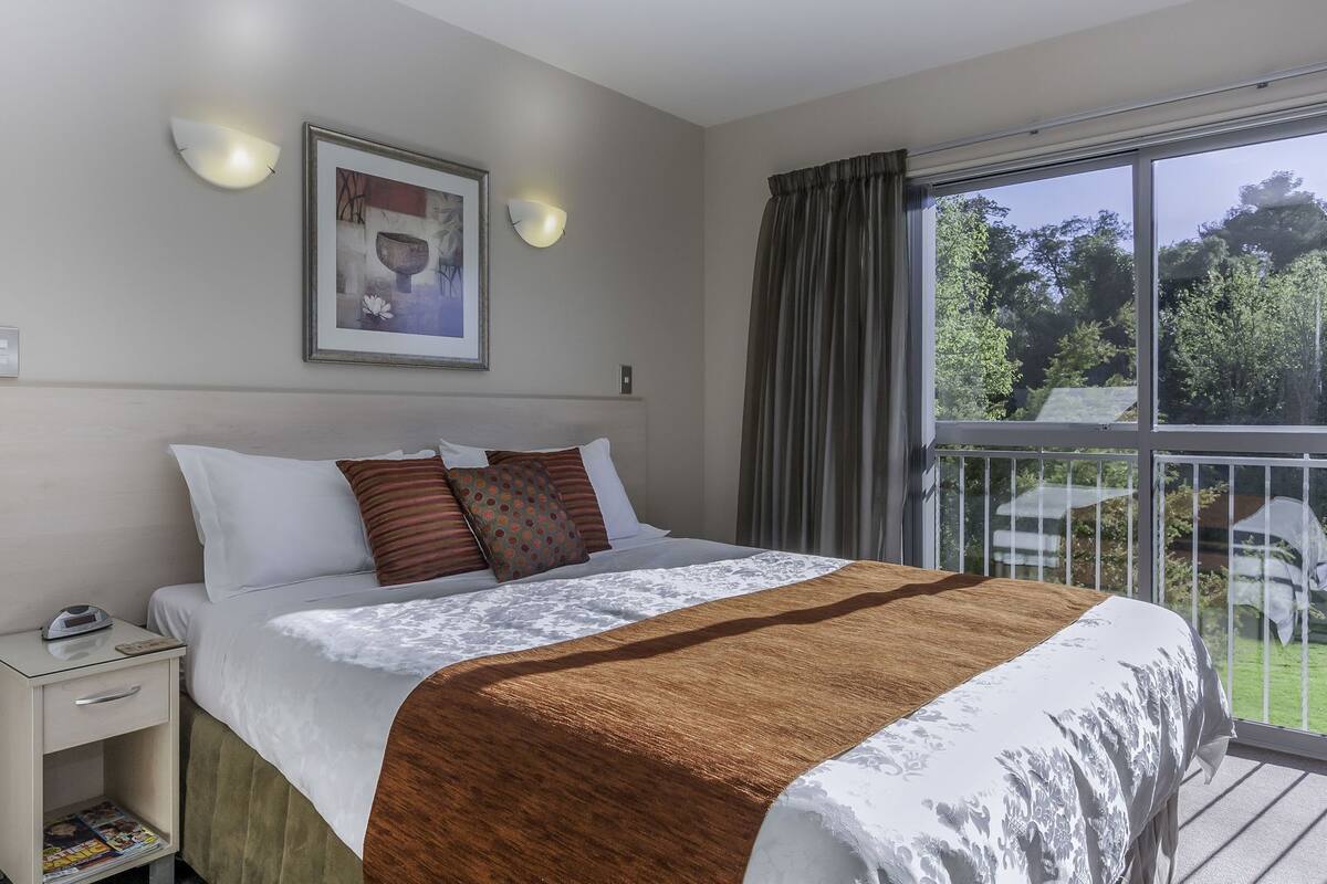 Een slaapkamer met een bed, nachtkastje, lamp, kunstwerk in een lijst en een balkon met uitzicht.