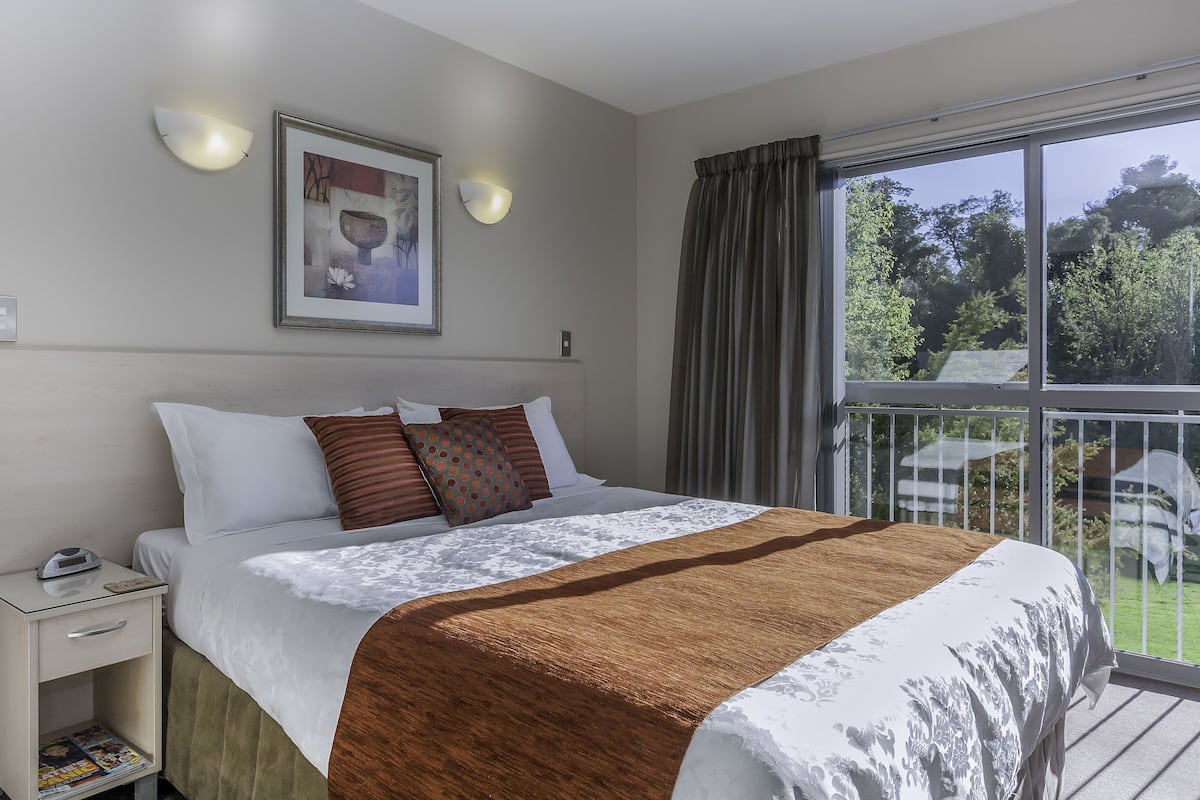 Een slaapkamer met een bed, nachtkastje, lamp en een raam met uitzicht op groen.