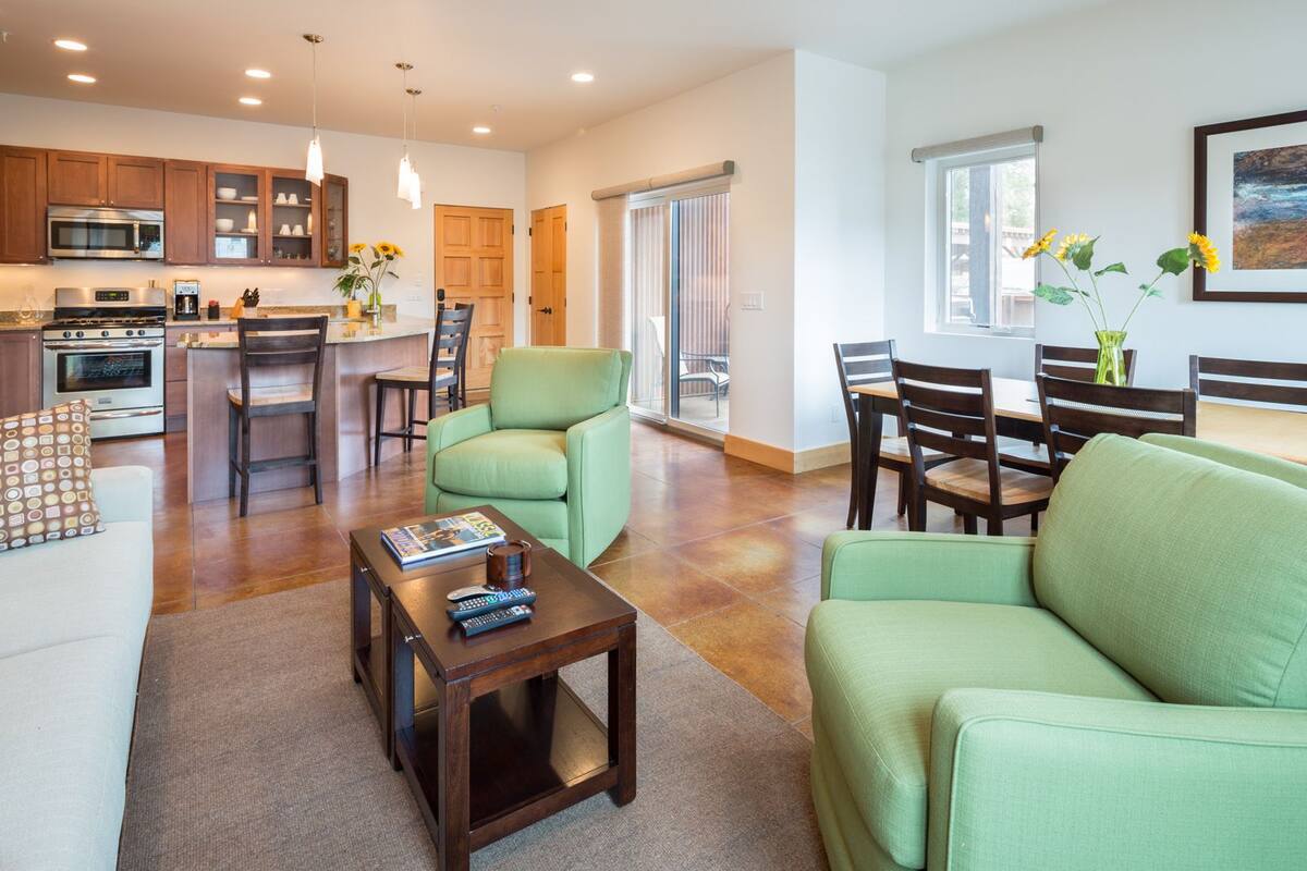 Ein modernes Wohnzimmer mit einem grünen Sofa, einem Couchtisch und einem Essbereich mit Holztisch und Stühlen. Es gibt eine Küchenzeile mit einer Mikrowelle und einer Kaffeemaschine.