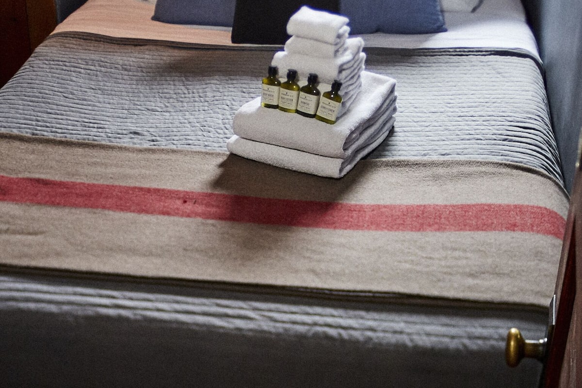 A neatly made bed with a striped blanket, pillows, and a stack of folded towels with bottles.