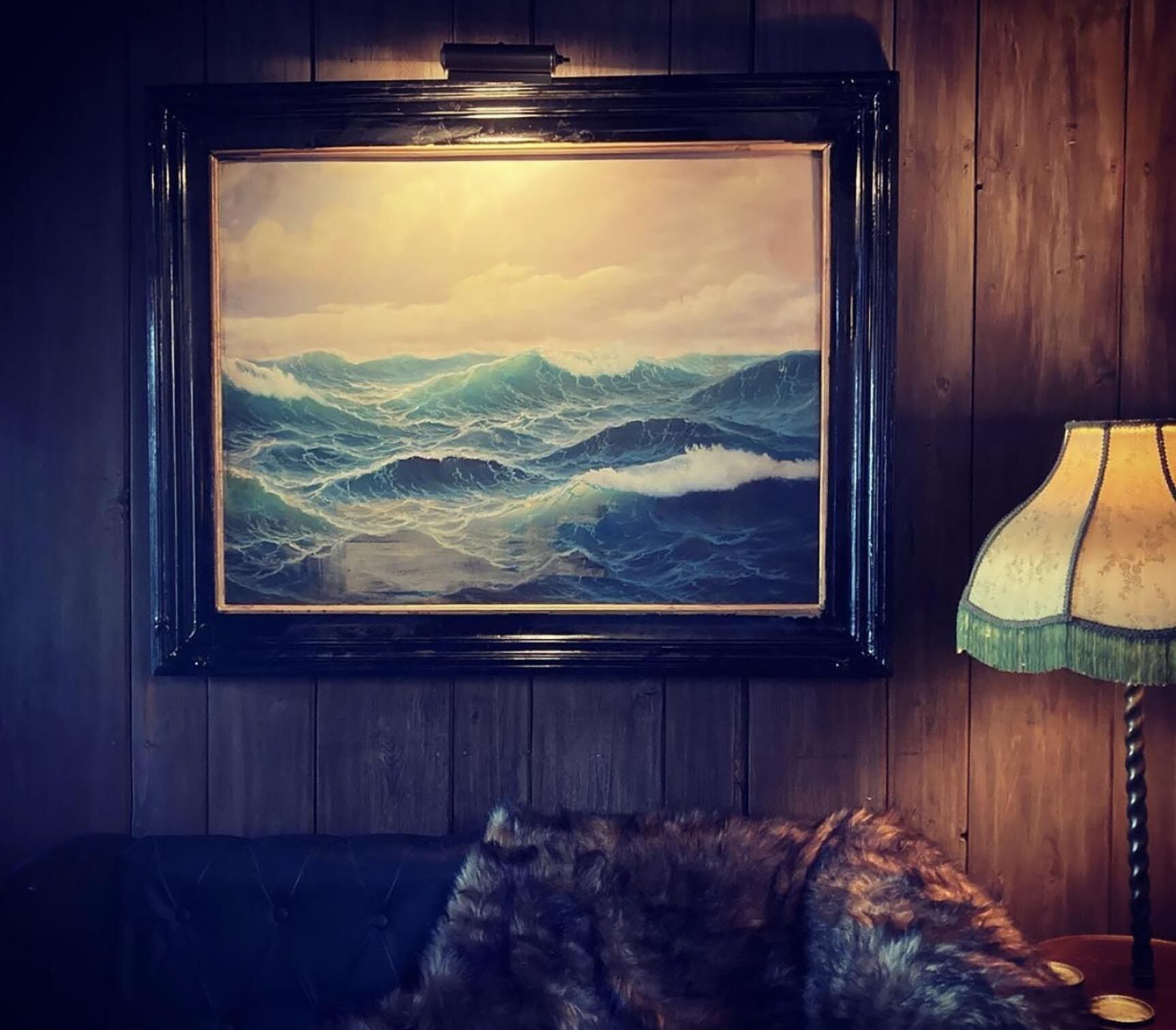 Un cuadro enmarco con mares embravecidos sobre una pared de madera, una lámpara y un sillón cubierto de piel.