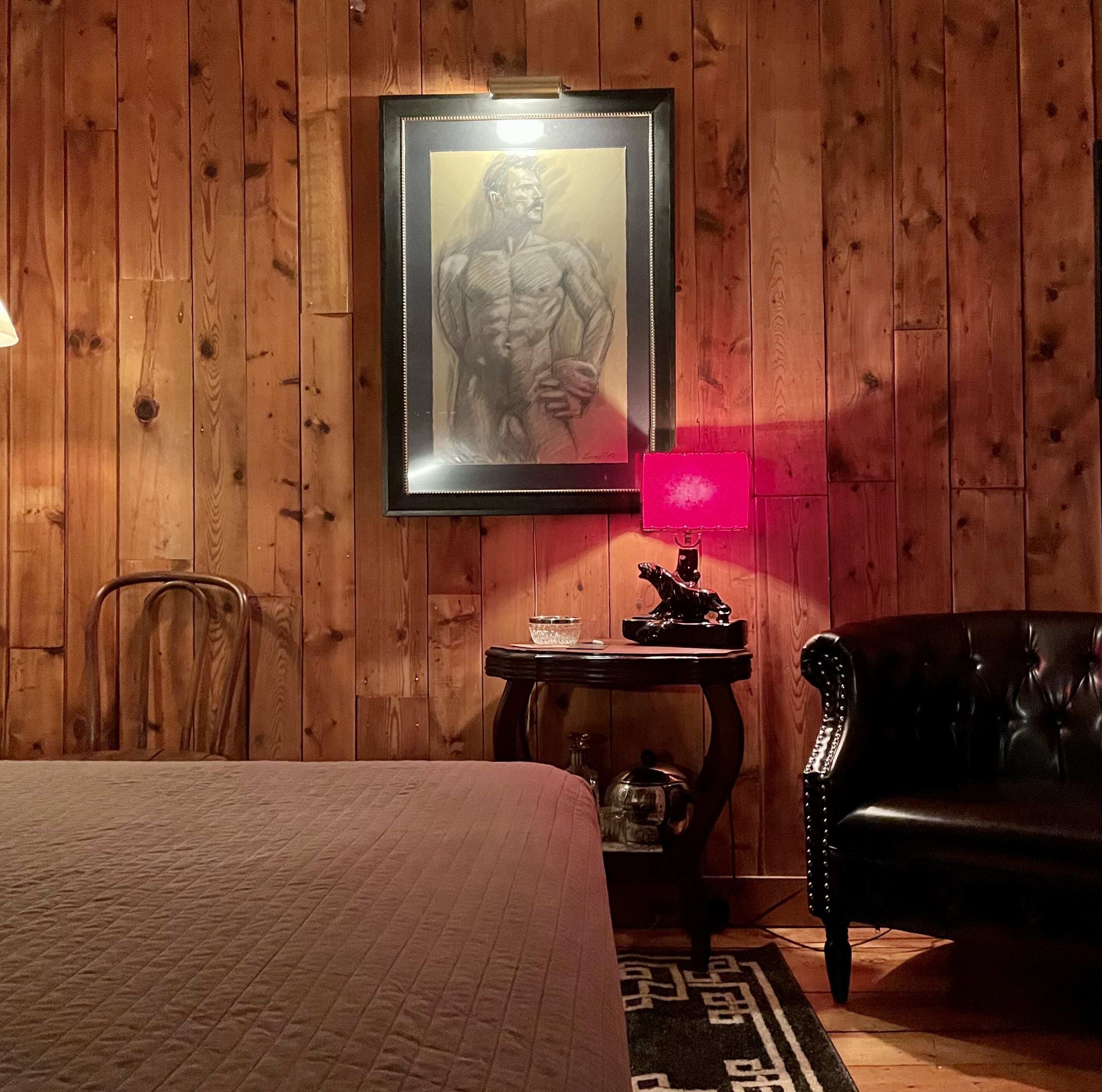 Une pièce aux murs en bois, avec un lit, un canapé en cuir noir, une petite table avec une lampe et un tableau encadré au mur.