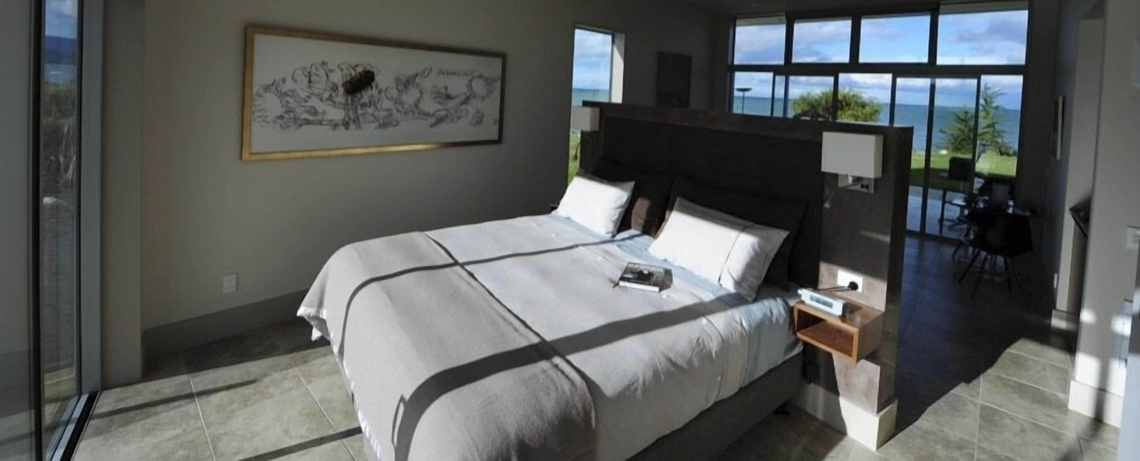 A modern bedroom with a large bed, a nightstand, a wall-mounted lamp, and a view of the ocean.