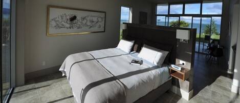 A modern bedroom with a large bed, a nightstand, a wall-mounted lamp, and a view of the ocean.
