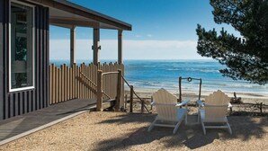 Une terrasse en bois mène à une maison de plage où se trouvent deux fauteuils Adirondack, tous deux orientés vers l’océan.