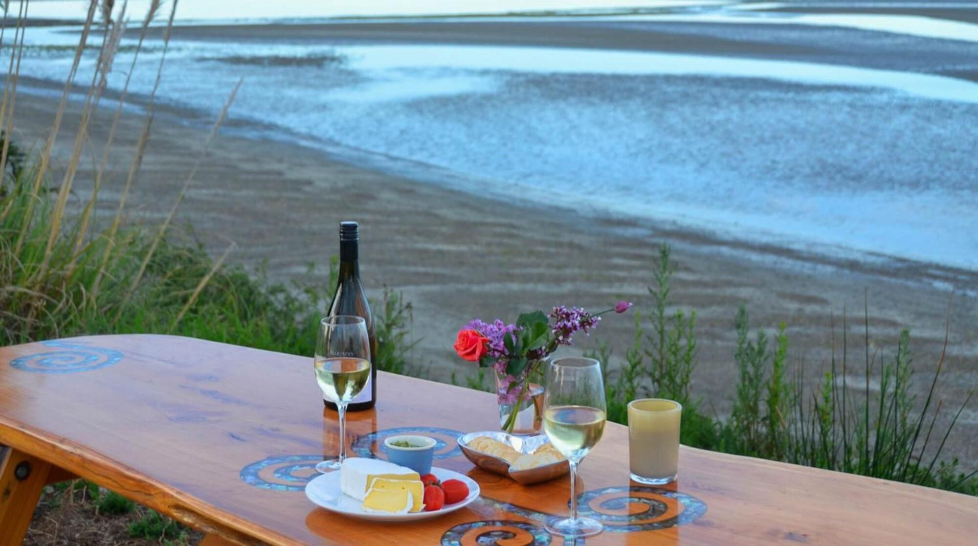 A wooden table with a bottle of white wine, a glass of white wine, a glass of milk, a plate with cheese and tomatoes, and a small vase with flowers.