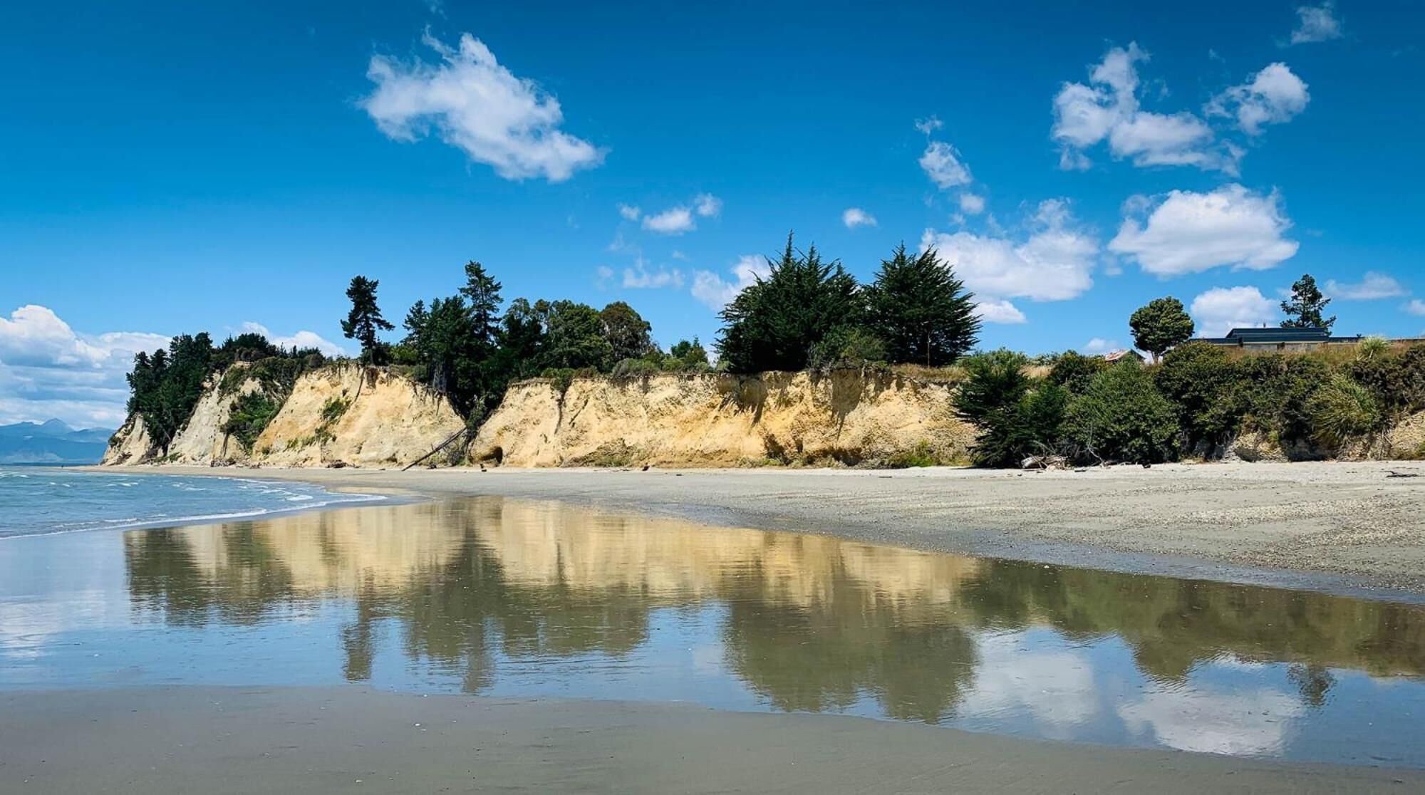 A beach with a clear reflection in the water, sandy shore, and a cliff with trees.