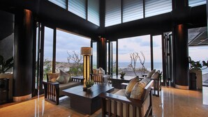 Un espace intérieur vaste et lumineux, avec une grande fenêtre donnant sur la mer, un ensemble de meubles de salon modernes et un grand lampadaire.