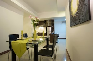 A dining area with a glass table, chairs, and a vase with flowers. There is a kitchen in the background with cabinets and appliances.