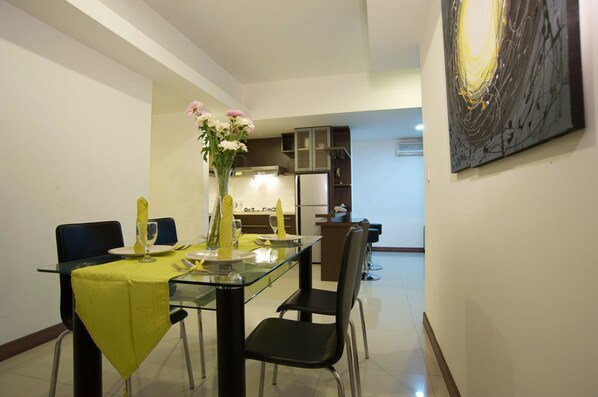 Un comedor con una mesa de vidrio, sillas y un florero con flores. Se observa una cocina al fondo con armarios y electrodomésticos.