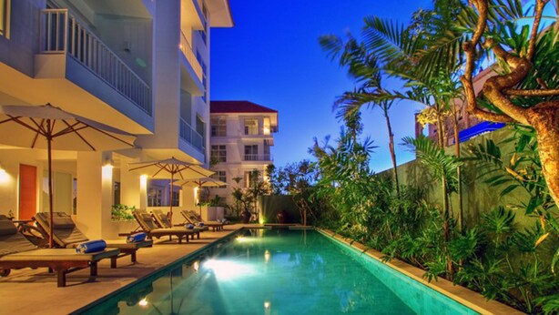 Piscina com espreguiçadeiras e guarda-sóis, cercada por vegetação e um edifício com varandas.