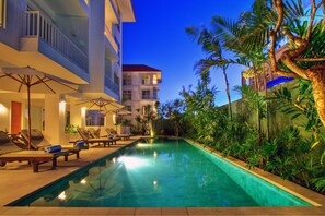Piscina con sillones de descanso y sombrillas, rodeada de vegetación y un edificio con balcones.