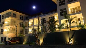 Um hotel de vários andares com varandas, um carro estacionado em frente e um céu noturno claro com a lua visível.